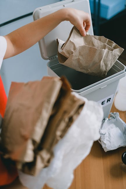 A person is disposing of crumpled brown paper bags into a white rubbish bin that features a lid slightly open, revealing more paper waste inside. The person's arm, visible in the image, is reaching to