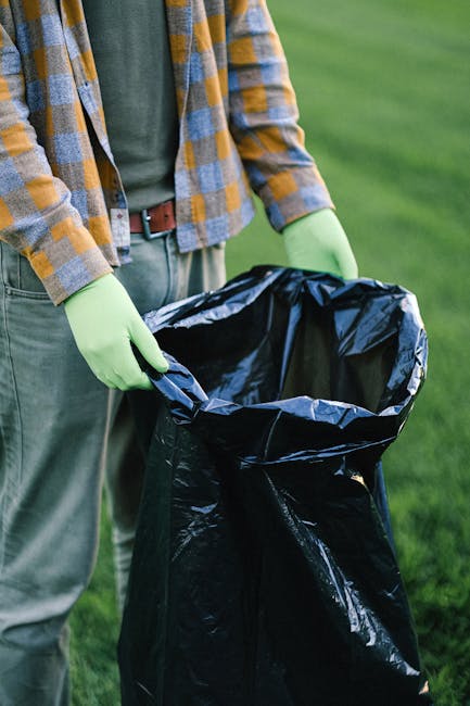 A narrow urban alleyway during daytime filled with accumulated waste and discarded debris, including large black garbage bags piled up towards the middle and back. The foreground features a wheeled tr