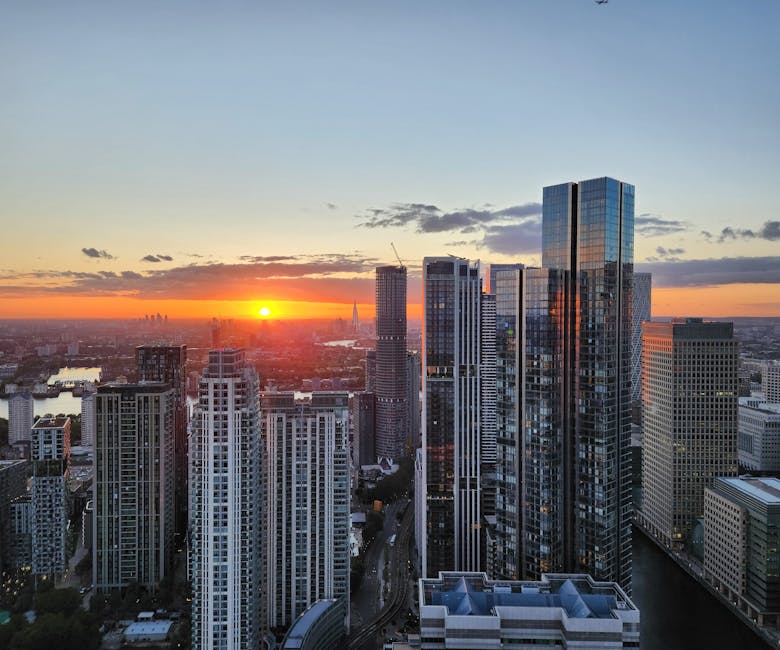 A panoramic view of a modern city skyline at sunset, featuring tall glass-clad skyscrapers with reflective surfaces capturing the orange and blue hues of the sky. In the foreground, a cluster of high-