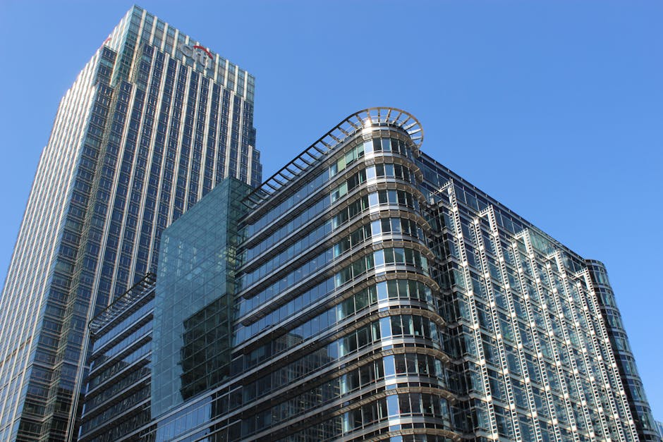 A panoramic view of a modern city skyline at sunset, featuring tall glass-clad skyscrapers with reflective surfaces capturing the orange and blue hues of the sky. In the foreground, a cluster of high-