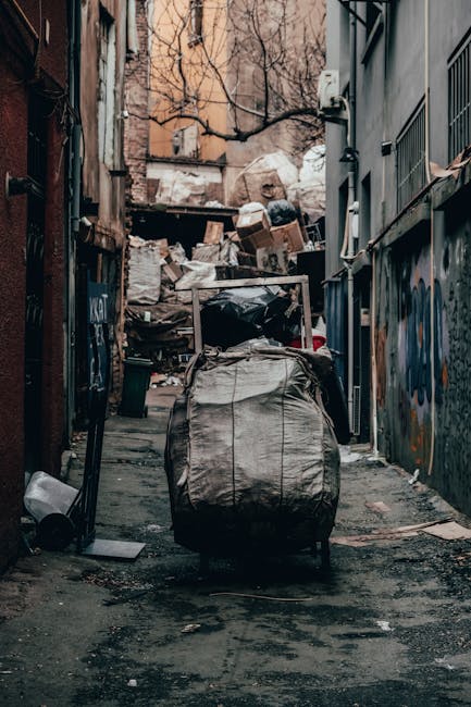 A narrow urban alleyway cluttered with a large, grey, industrial-looking garbage bag in the foreground, positioned on the asphalt surface. Behind it, there is a significant accumulation of mixed rubbi