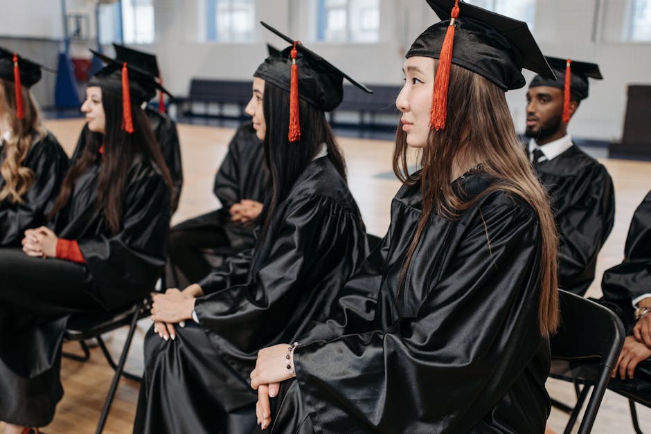 Three young women dressed in black graduation gowns and mortarboards are engaged in a celebratory moment outdoors. The woman on the right, with long brown hair and a bright smile, is handing a rolled